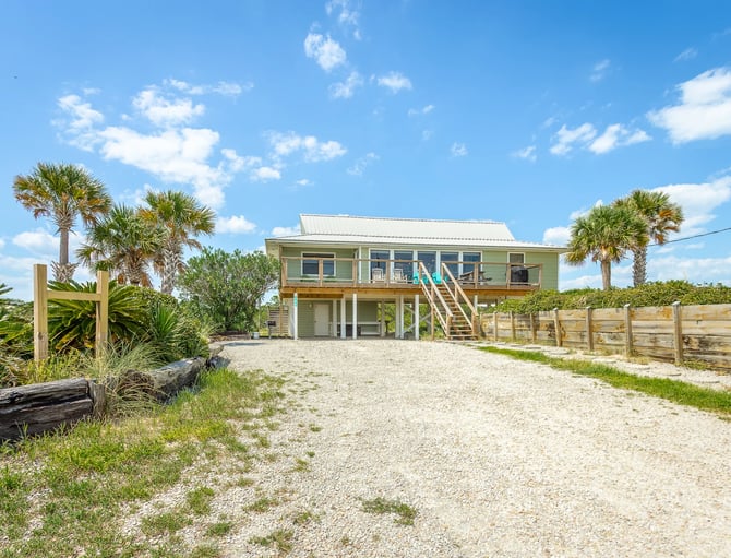 Elevated beach house with green exterior, wooden deck, and palm trees on sandy lot with dune fencing